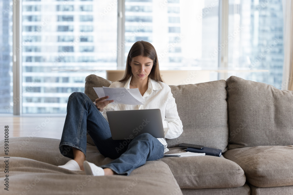 Busy woman sitting on couch with laptop on laps holding bills, makes easy remote e-payment through e-banking application, student prepare for college exams, studying at home using internet connection Busy woman sitting on couch with laptop on laps holding bills, makes easy remote e-payment through e-banking application, student prepare for college exams, studying at home using internet connection
