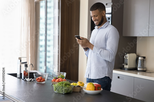 Young handsome African guy using cellphone standing in modern kitchen, search vegetarian recipe on-line cookbook, using calories counter while cooking healthy dish for weigh loss. Modern tech, cuisine