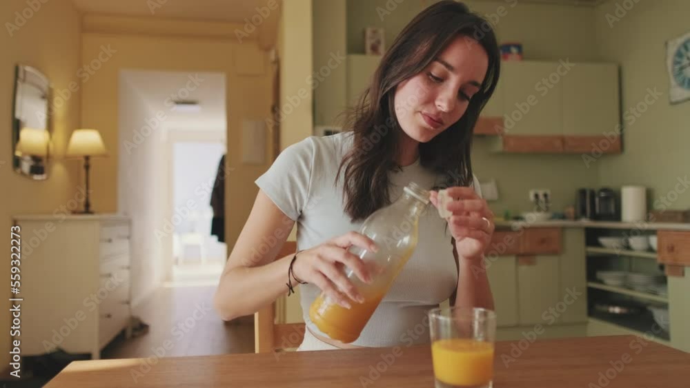 Young woman pouring orange juice into glass while sitting in the kitchen Young woman pouring orange juice into glass while sitting in the kitchen