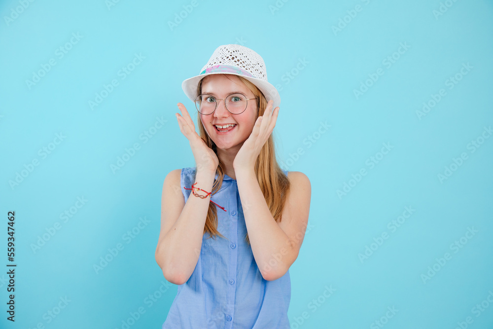 Cheerful teen girl stand with happy emotion on empty blue background. Portrait of smiling school aged child in stylish sleeveless shirt, glasses and cute panama. Teenage fashion, summer wear Cheerful teen girl stand with happy emotion on empty blue background. Portrait of smiling school aged child in stylish sleeveless shirt, glasses and cute panama. Teenage fashion, summer wear