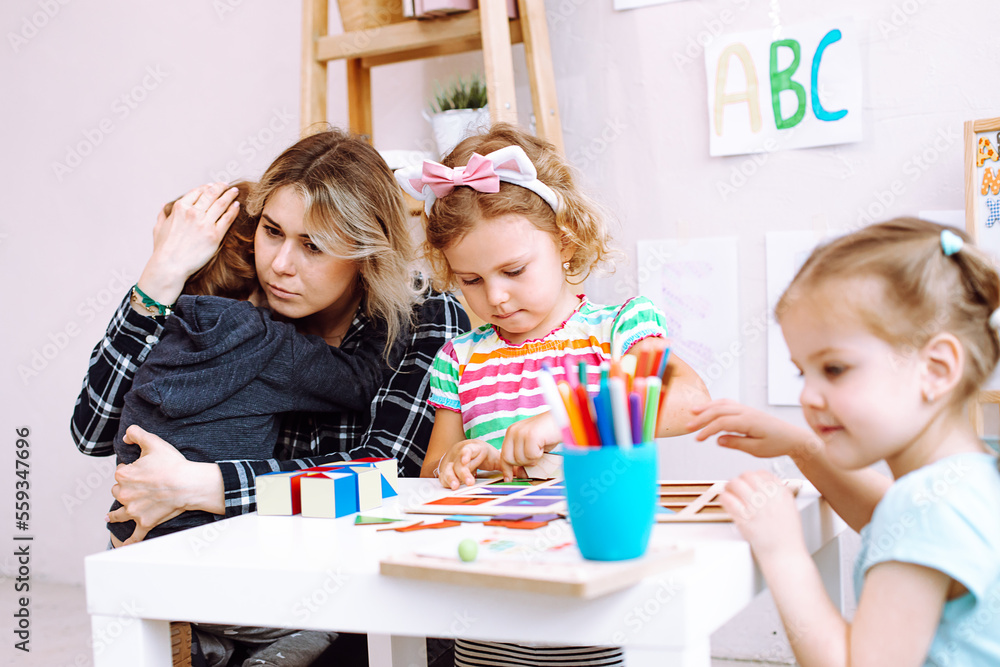 Fototapeta premium Young educator embrace and calm down crying boy in kindergarten. Pretty little girls with interest playing with educational toys on desk. Fail, support, hugs, lesson in daycare centre