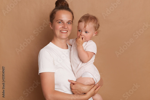 Portrait of joyful woman with bun hairstyle wearing white T- shirt standing with her little daughter in hands isolated over brown background, looking at camera with toothy smile.