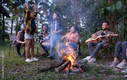 Group of family camper van sitting by fire grilling sausages and singing dancing have fun