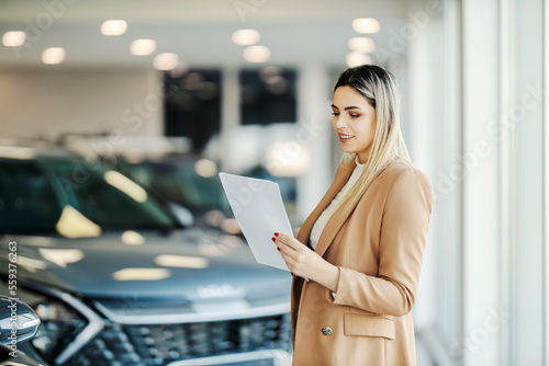 A happy female car dealer is standing in showroom with paperwork in her hands and looking at it.