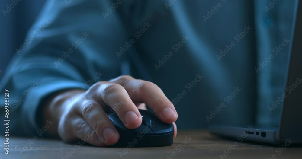Close up man's hand uses a computer mouse while working on a laptop in the evening. Male freelancer clicking black mouse while browsing internet or working on computer remotely from home office.
