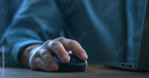 Close up man's hand uses a computer mouse while working on a laptop in the evening. Male freelancer clicking black mouse while browsing internet or working on computer remotely from home office. photo