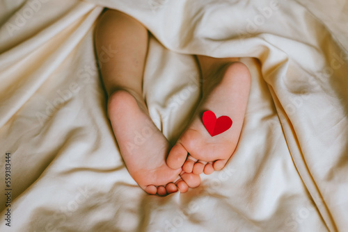 Children's feet and heels with heart on a white bed to Christmas, Valentine's day, Mother's Day. Infant baby is sleeping in his crib. Importance of sleep for babies. Sleep mode babies 3 y.o.