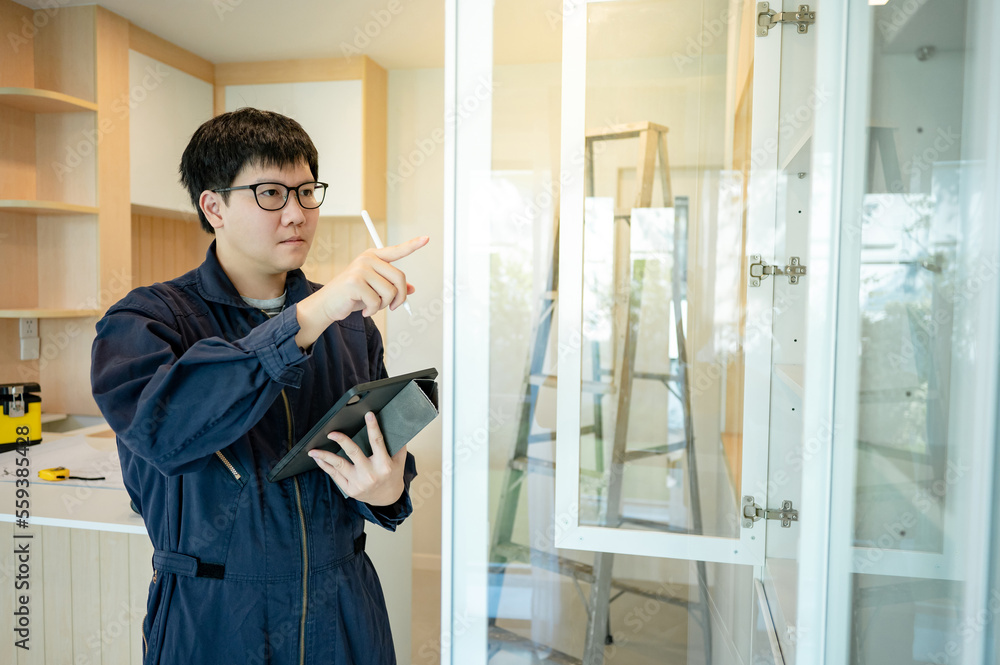 Home renovation or House remodeling concept. Asian male interior construction worker or furniture assembler man using digital tablet checking glass panel of cabinet installation.