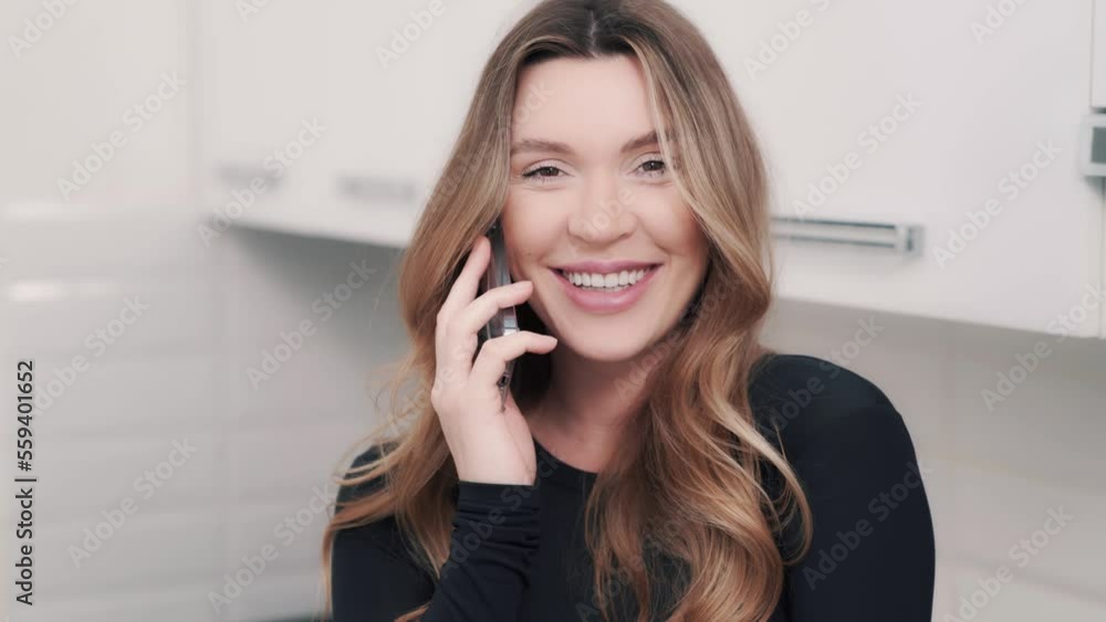 Indoor portrait of a charming woman with long dark blond hair talking on the phone with a happy look. Girl laughing during the friendly conversation and looking into the camera Indoor portrait of a charming woman with long dark blond hair talking on the phone with a happy look. Girl laughing during the friendly conversation and looking into the camera