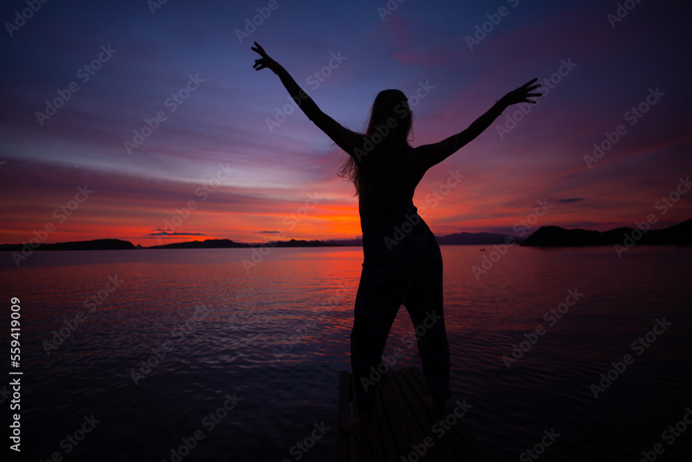 Girl With Arms Up Enjoys Colorful Sunset In Komodo Islands, Indonesia Girl With Arms Up Enjoys Colorful Sunset In Komodo Islands, Indonesia