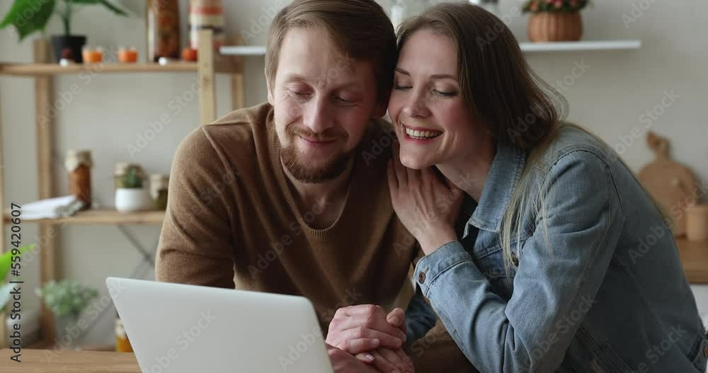 Close up smiling couple in love use laptop seated in domestic kitchen, staring at screen discuss purchase, buy gifts, order foods via on-line supermarket, talking, enjoy easy comfort retail services Close up smiling couple in love use laptop seated in domestic kitchen, staring at screen discuss purchase, buy gifts, order foods via on-line supermarket, talking, enjoy easy comfort retail services