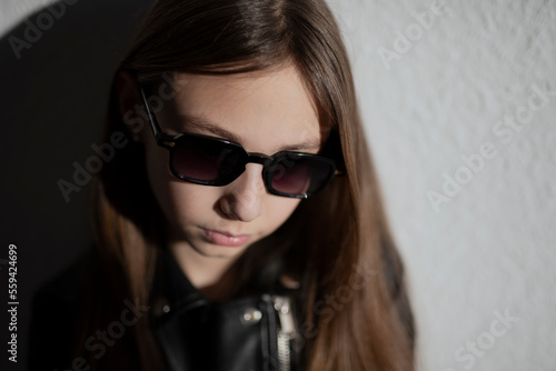 portrait of a stylish child in leather jacket with glasses on dark background with a shadows © Alona
