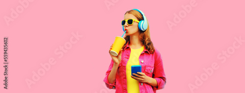 Portrait of stylish young woman listening to music in headphones with cup of fresh juice wearing jacket on pink background