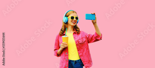 Portrait of happy smiling woman taking selfie with smartphone listening to music in wireless headphones wearing jacket on vivid pink background