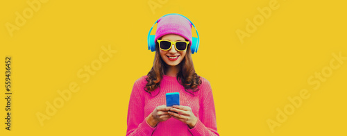 Portrait of modern young woman in wireless headphones listening to music with smartphone wearing knitted sweater, pink hat on yellow background