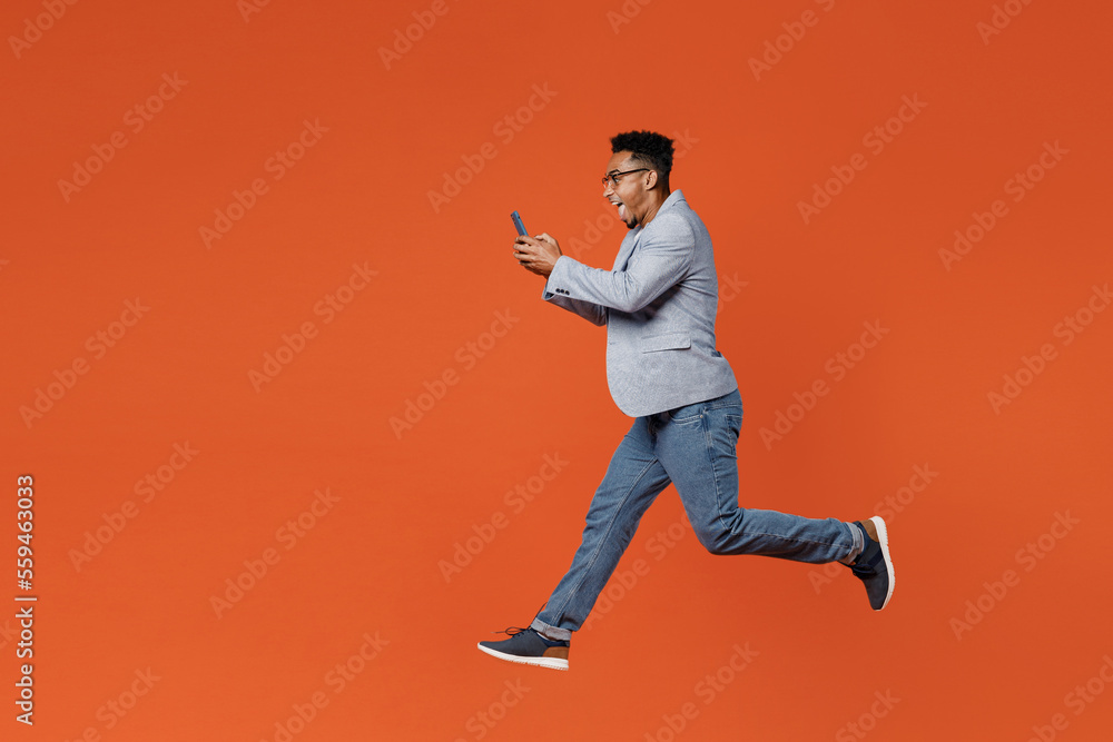 Full body young employee business man corporate lawyer wear classic formal grey suit shirt glasses work in office jump high hold in hand use mobile cell phone isolated on plain red orange background. Full body young employee business man corporate lawyer wear classic formal grey suit shirt glasses work in office jump high hold in hand use mobile cell phone isolated on plain red orange background.