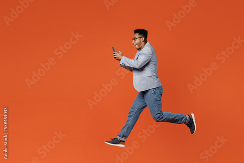 Full body young employee business man corporate lawyer wear classic formal grey suit shirt glasses work in office jump high hold in hand use mobile cell phone isolated on plain red orange background.