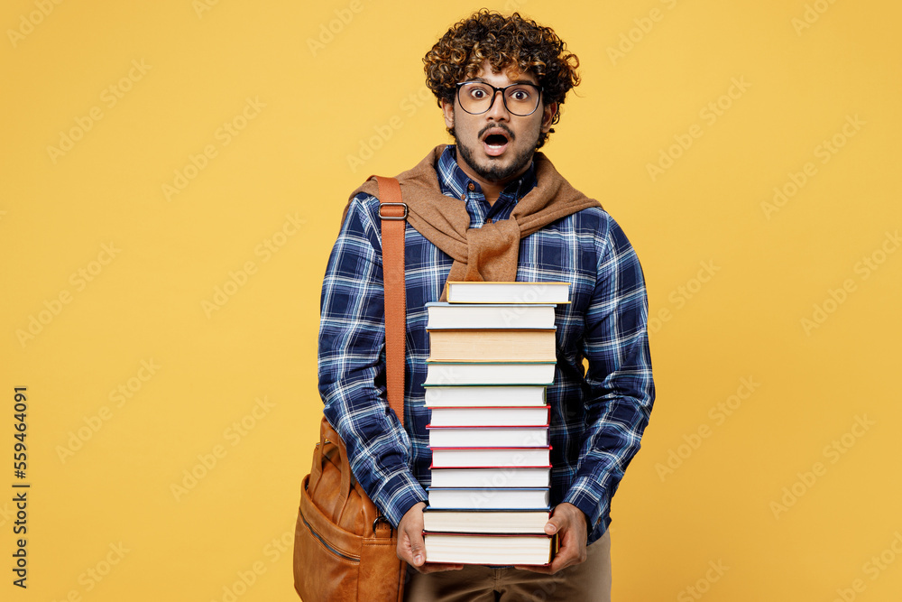 Young shocked surprised teen Indian boy IT student wear casual clothes shirt glasses bag hold in hands pile of books isolated on plain yellow color background High school university college concept - obrazy, fototapety, plakaty 