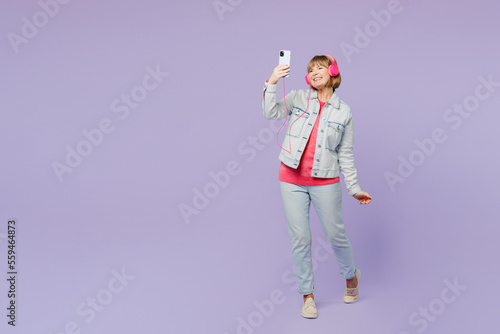 Full body elderly fun blonde woman 50s years old wears casual clothes denim jacket t-shirt listen to music in headphones use mobile cell phone dance isolated on plain pastel light purple background.