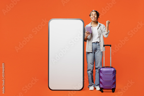 Full body traveler woman in grey shirt near big blank screen mobile cell phone bag hold pasport ticket do winner gesture isolated on plain orange background Tourist travel in free time rest getaway.