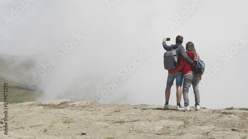 Couple standing with back on mountain peak, taking pictures cloudy sky
