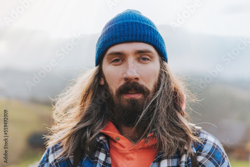 Bild auf Leinwand Portrait of attractive man with long hair wearing blue hat and jacket with mountain on the background