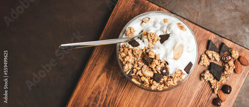 Granola cereal oatmeal with white yogurt, chocolate, fruist and nuts in a bowl on dark wooden board, top view