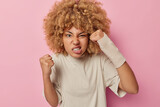 Photo of angry woman frowns face looks angrily at you clenches fists wears bandage on arm has injured hand dressed in casual beige t shirt isolated over pink background. Wrist pain health care concept