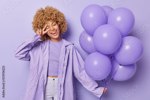 Horizontal shot of positive woman with curly hair makes peace sign holds inflated balloons gathered in bunch dressed in outerwear poses against purple background celebrates special occasion. © wayhome.studio 