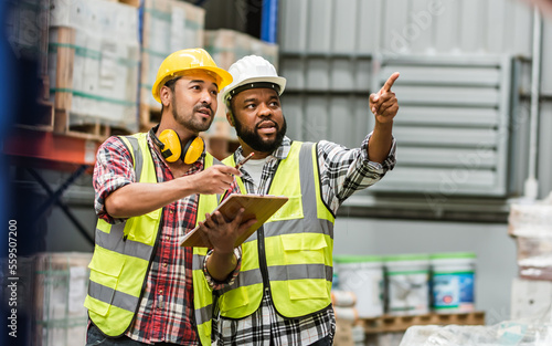 Photo Two diverse professional male workers holding board, checking shipping stocks in storage, warehouse or factory for delivery, wearing safety hat