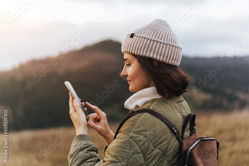 Happy smiling female traveller using cell telephone with online map and texting to friends, blurred forest and mountains in the background, concept of using technologies in nature
