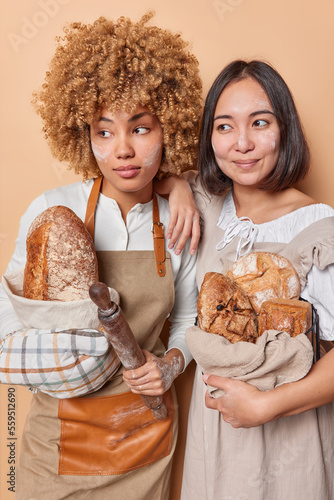 Vertical shot of pensive female bakers stand next to each other dressed in uniform hold crusty homemade loaves of bread isolated over brown studio background. Healthy organic food and bakery concept