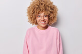 Portrait of positive curly haired woman bites lips looks gladfully at camera hears something has curious expression wears big transparent eyeglasses and pink pullover isolated over grey background