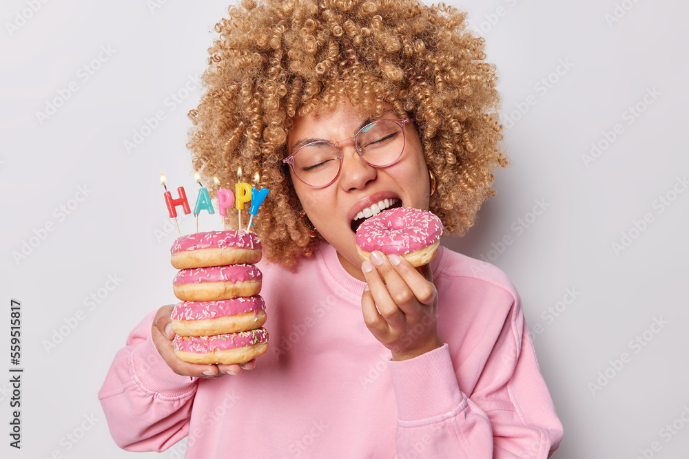 Photo of curly haired adult woman bites appetizing glazed doughnut holds pile of donuts with burning candles eats harmful food wears spectacles and pink pullover isolated over grey background
