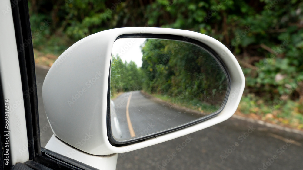 Mirror view of white car travel on the asphalt road. with blurred of green forest beside road at day after rainy.