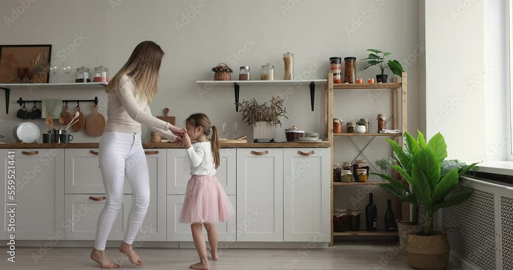 Happy young mother hold hands dancing with preschooler adorable daughter in festive fluffy pink skirt, moving to music barefoot in modern warm domestic kitchen, having fun on weekend celebrate event Happy young mother hold hands dancing with preschooler adorable daughter in festive fluffy pink skirt, moving to music barefoot in modern warm domestic kitchen, having fun on weekend celebrate event