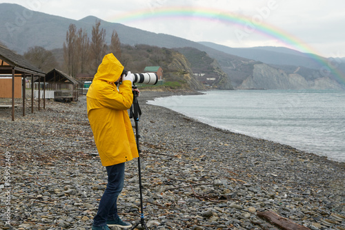 A man photographs a rainbow on the beach with a long-focus lens resting on a monopod