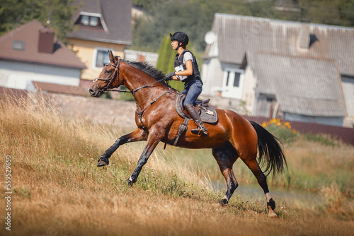 portrait of attractive young rider woman and bay mare horse galloping during eventing cross country competition in summer