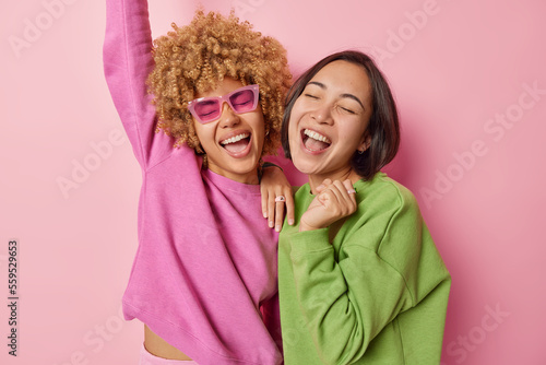 Horizontal shot of two women express positivity and joy laugh happily and exclaim loudly stand closely dressed in casual jumpers stand pleased isolated over pink background. Friendship and emotions