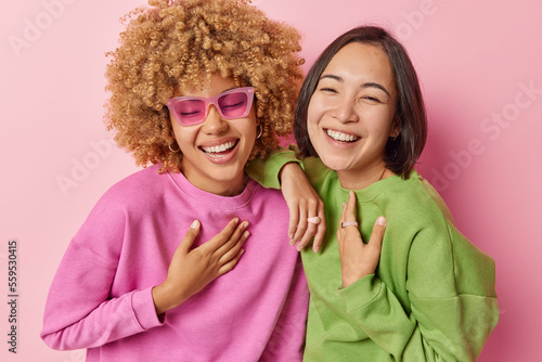 Принти на полотні Overjoyed multiracial women laugh happily smile toothily keep hands on chest have upbeat mood wear sunglasses casual jumpers stand closely to each other isolated on pink background