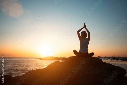 A woman does yoga in the lotus pose, meditating on the rocks of the Atlantic during sunset.