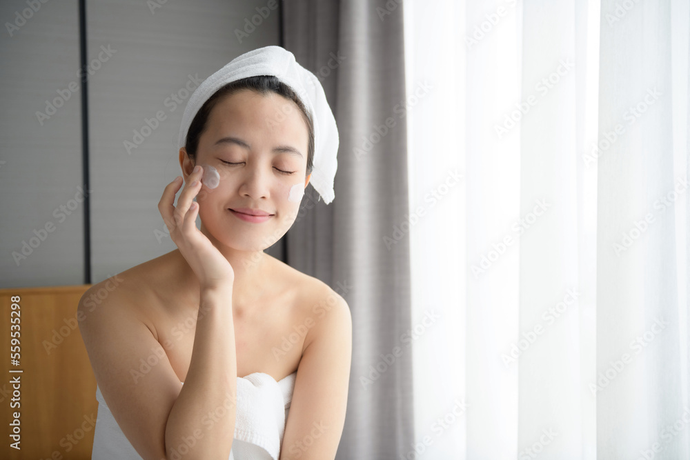Happy young Asian woman applying face lotions while wearing a towel and touching her face. Daily makeup and skincare Happy young Asian woman applying face lotions while wearing a towel and touching her face. Daily makeup and skincare