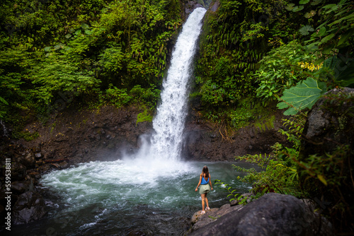 Fotografia, Obraz A beautiful girl stands on rocks under a powerful tropical waterfall in Costa Ri
