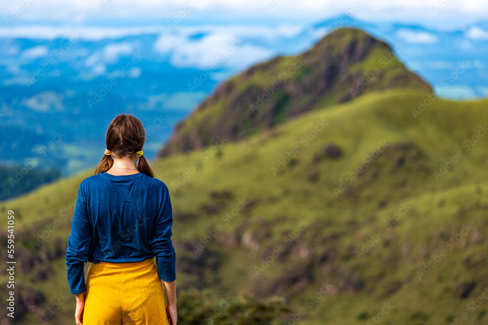A backpacker girl stands on top of Costa Rica's cerro pelado mountains during a sunny day; hiking through mighty mountains covered with green succulent grass; mountains in the tropics amid rainforests