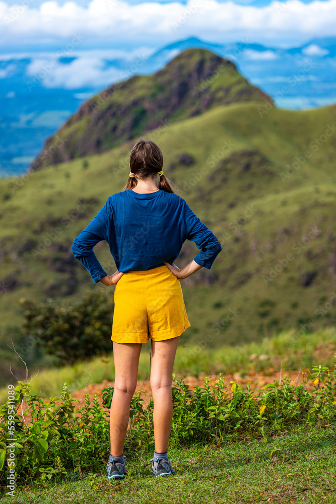 A backpacker girl stands on top of Costa Rica's cerro pelado mountains during a sunny day; hiking through mighty mountains covered with green succulent grass; mountains in the tropics amid rainforests