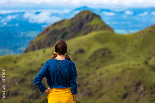 A backpacker girl stands on top of Costa Rica's cerro pelado mountains during a sunny day; hiking through mighty mountains covered with green succulent grass; mountains in the tropics amid rainforests
