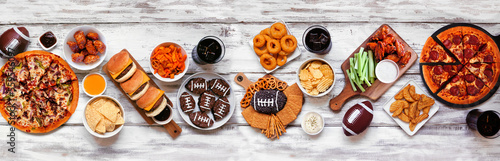 Super Bowl or football theme food table scene. Pizza, hamburgers, wings, snacks and sides. Top down view on a white wood background.