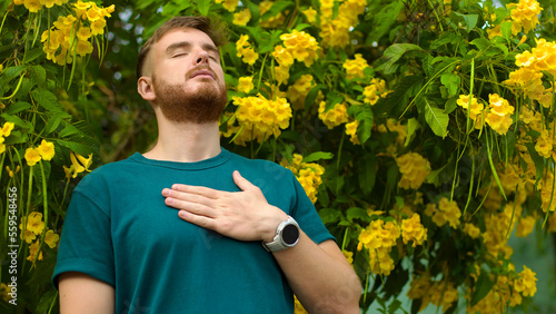 Portrait of happy handsome bearded guy, young positive man with beard is smelling beautiful yellow flowers in the garden, smiling, enjoying spring or summer day, breathing deep deeply fresh air