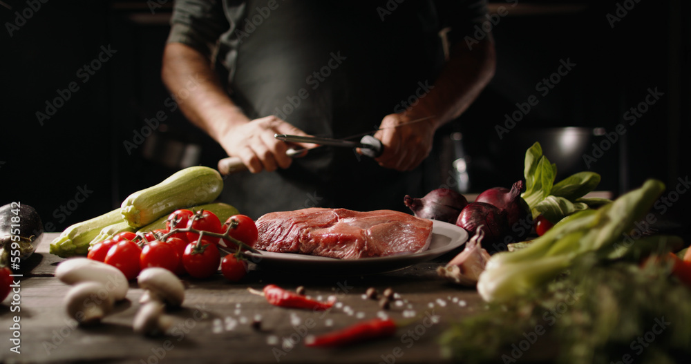 Chef sharpening his knife in front of kitchen table. Cooker preparing his tools before cutting raw piece of meat and various vegetables