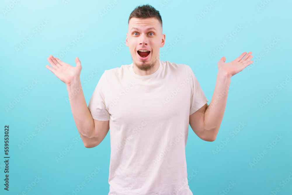 A сaucasian happy and surprised man in a white t-shirt looks at the camera spreading his hands to say wow on blue background. Outfit. Shocked. Modern. Style. Arm. Expression. Lifestyle. Overwhelmed A сaucasian happy and surprised man in a white t-shirt looks at the camera spreading his hands to say wow on blue background. Outfit. Shocked. Modern. Style. Arm. Expression. Lifestyle. Overwhelmed